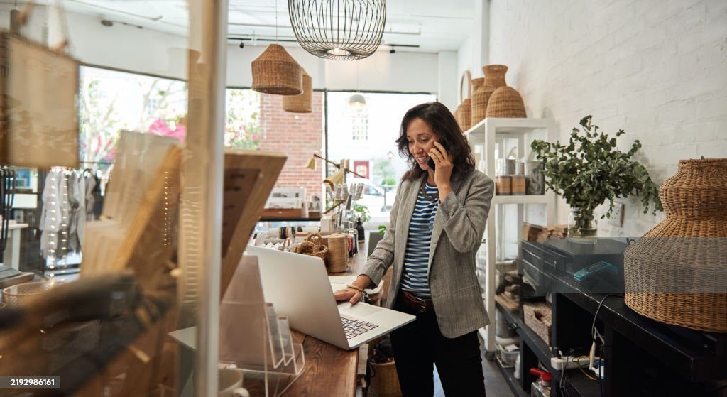 Shop owner talking on the phone while using a laptop at her store counter