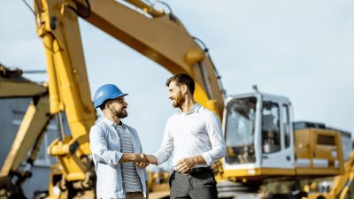 Two men shaking hands in front of an excavator at a construction site
