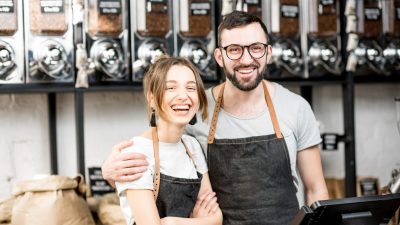 Two smiling small business owners in aprons standing in their bulk food shop