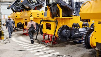 Factory workers walking past yellow heavy machinery on a manufacturing floor